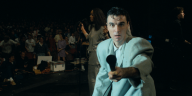 David Byrne, im hellen Anzug, hält auf der Bühne ein Mikrofon in die Kamera.